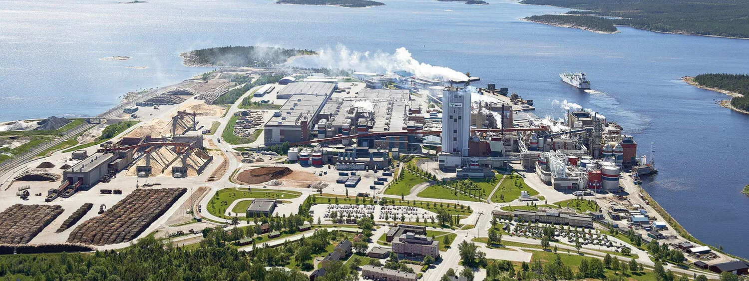 Aerial view of waterfront pulp mill with buildings, log stacks, smoke emissions, and a ship docked nearby