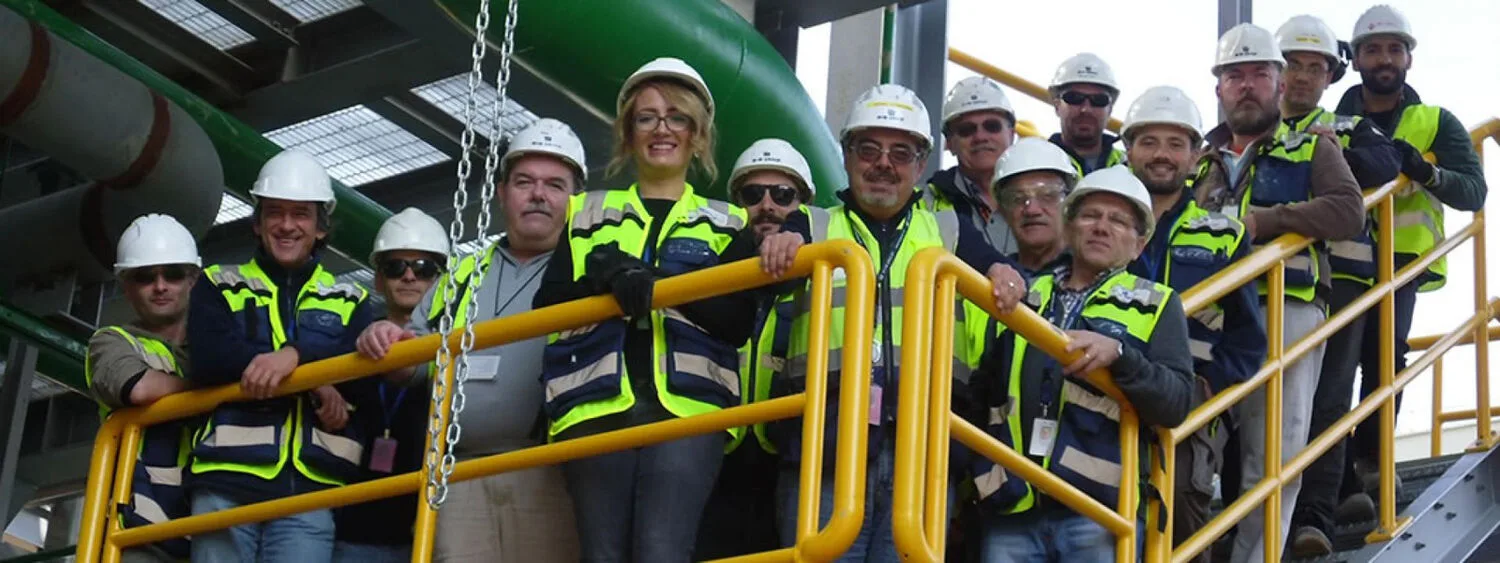 Group of people in safety helmets and high-visibility vests on yellow-railed platform in industrial setting with green pipes and metal structures
