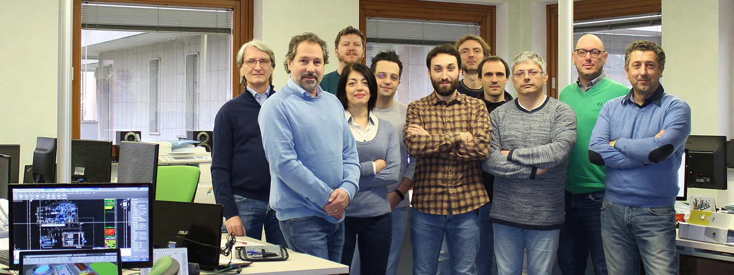 Group of eleven people in an office with computer monitors displaying technical diagrams, representing an engineering team at work