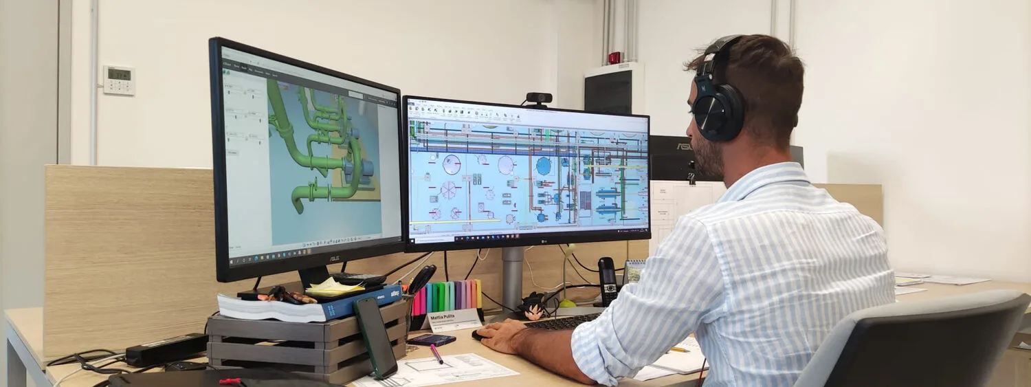 Person working at a desk with two monitors showing a 3D piping model and an engineering diagram, surrounded by office supplies
