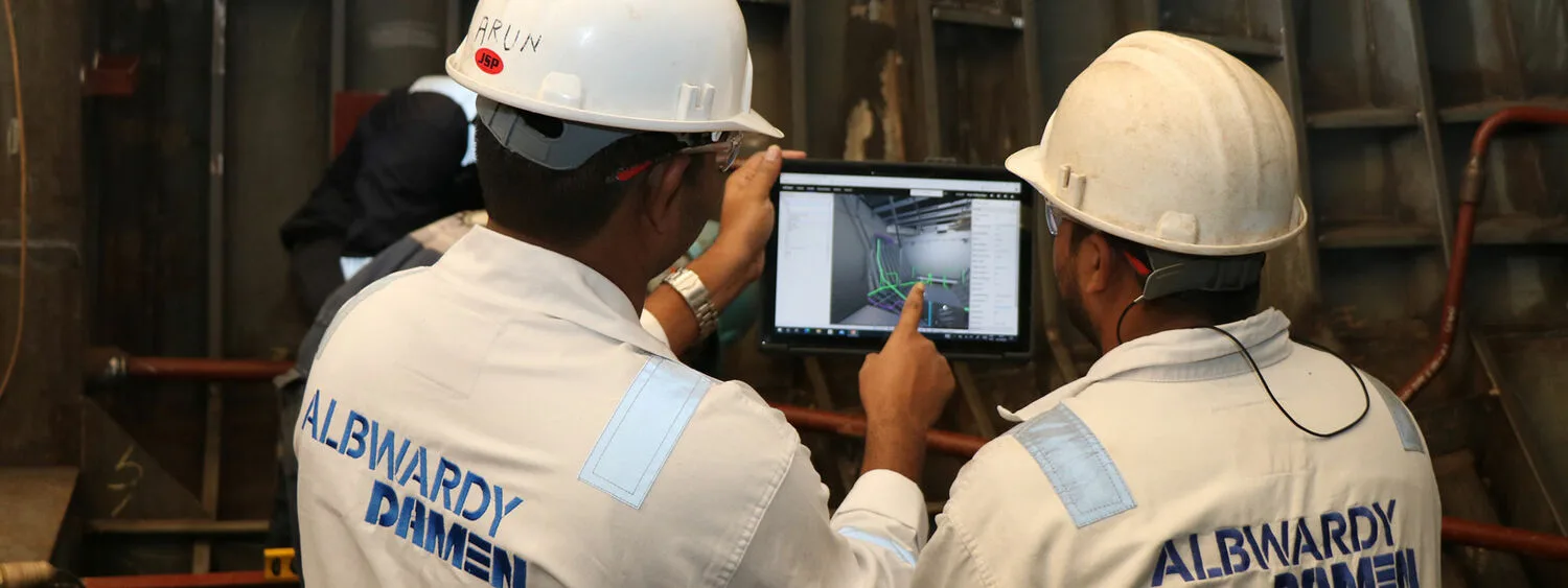 Two people in ALBWARDY DAMEN jackets and helmets reviewing a 3D model on a tablet in an industrial setting