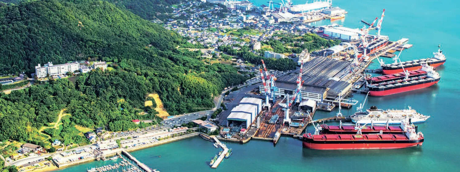 Aerial view of Tsuneishi shipyard with large docked vessels, cranes, industrial buildings, and green hills in the background.