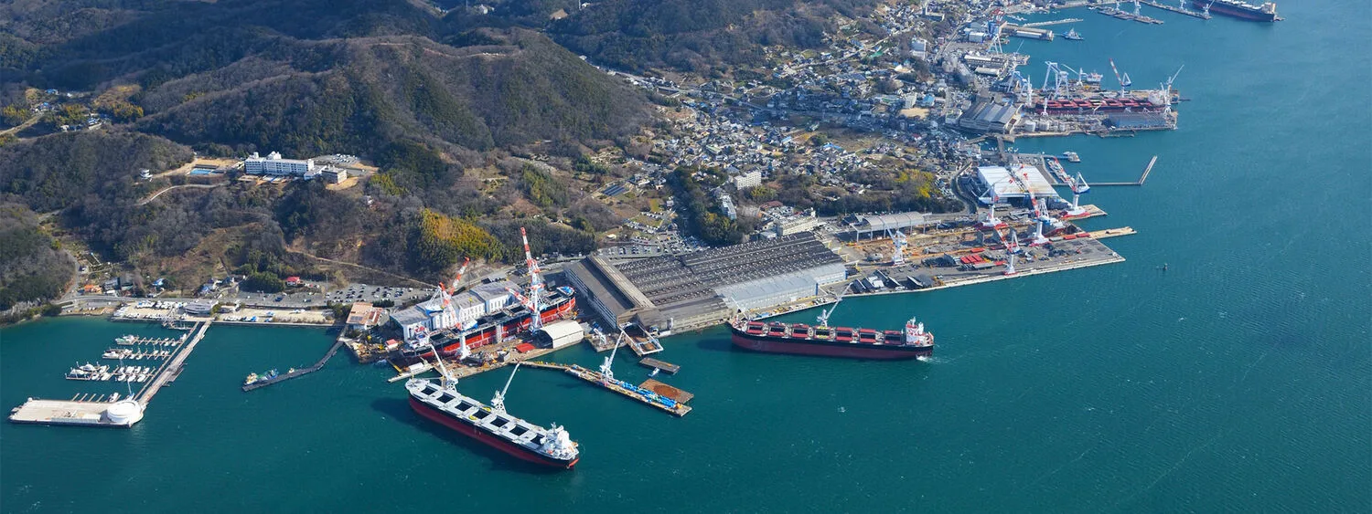 Aerial view of Tsuneishi shipyard with large docked vessels, cranes, industrial buildings, and surrounding urban and hilly landscape
