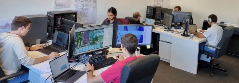 Six people working at desks with multiple monitors displaying CAD software in an office with posters and diagrams on the walls