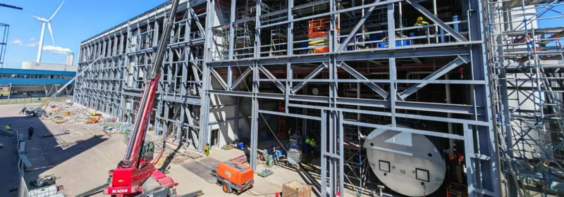 Industrial construction site with steel framework, red MAGNI crane, workers, and a wind turbine in the background