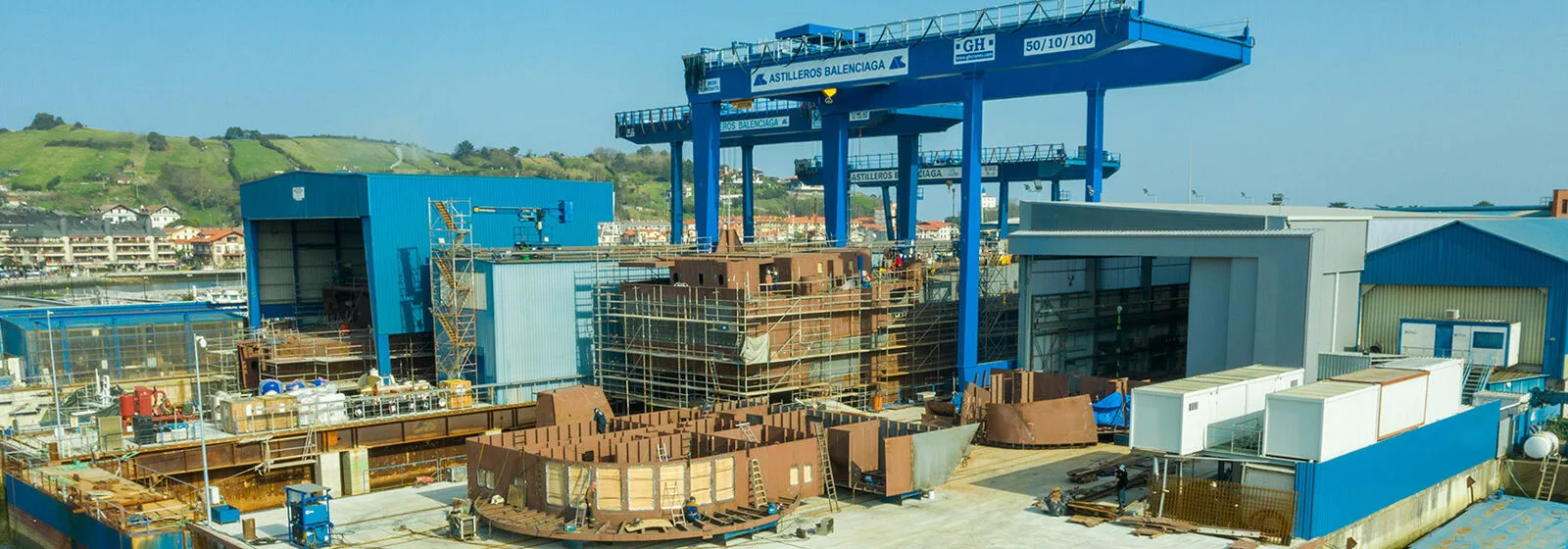 Shipyard with blue cranes labeled 'ASTILLEROS BALENCIAGA', ship hull sections under construction, and industrial buildings near green hills
