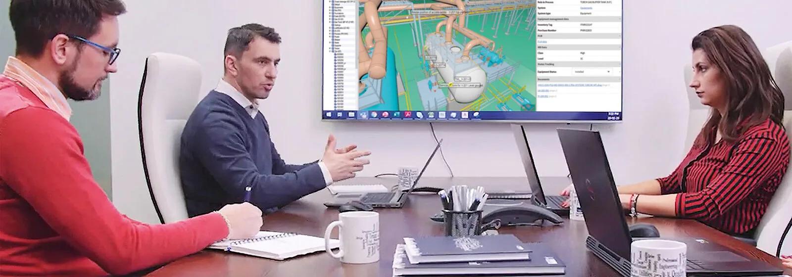 Three people in an office viewing a 3D industrial plant model on a large screen, with laptops and notebooks on the table