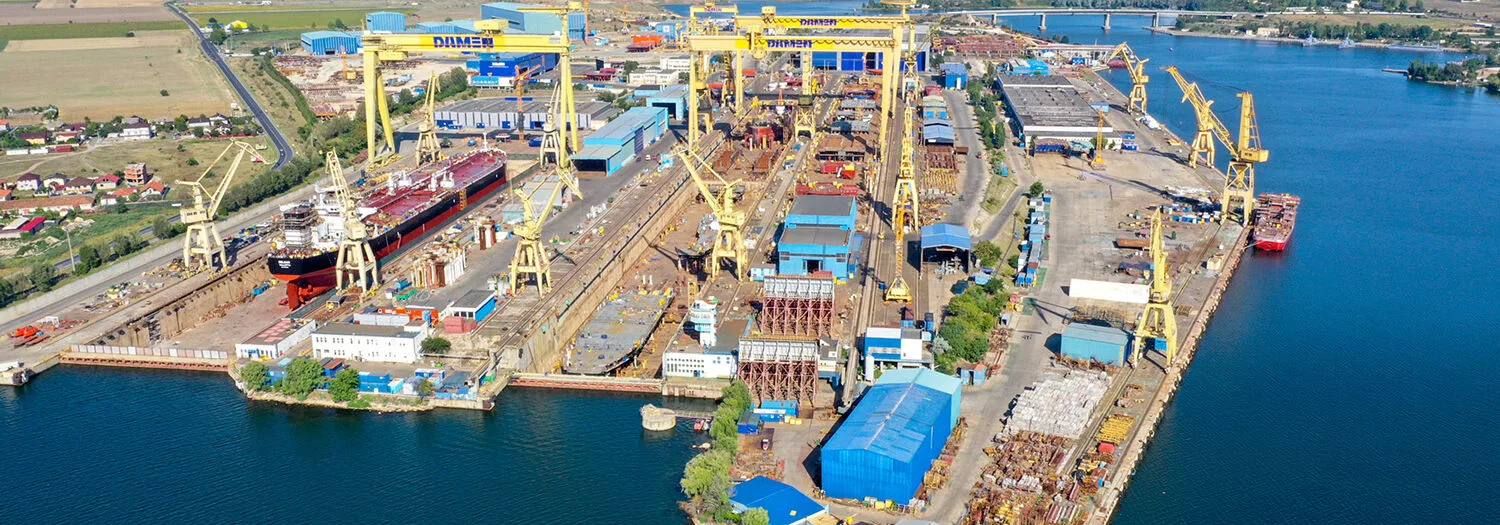 Aerial view of Damen Shipyards Mangalia with yellow cranes, dry docks, ships under construction, and industrial buildings by the water.