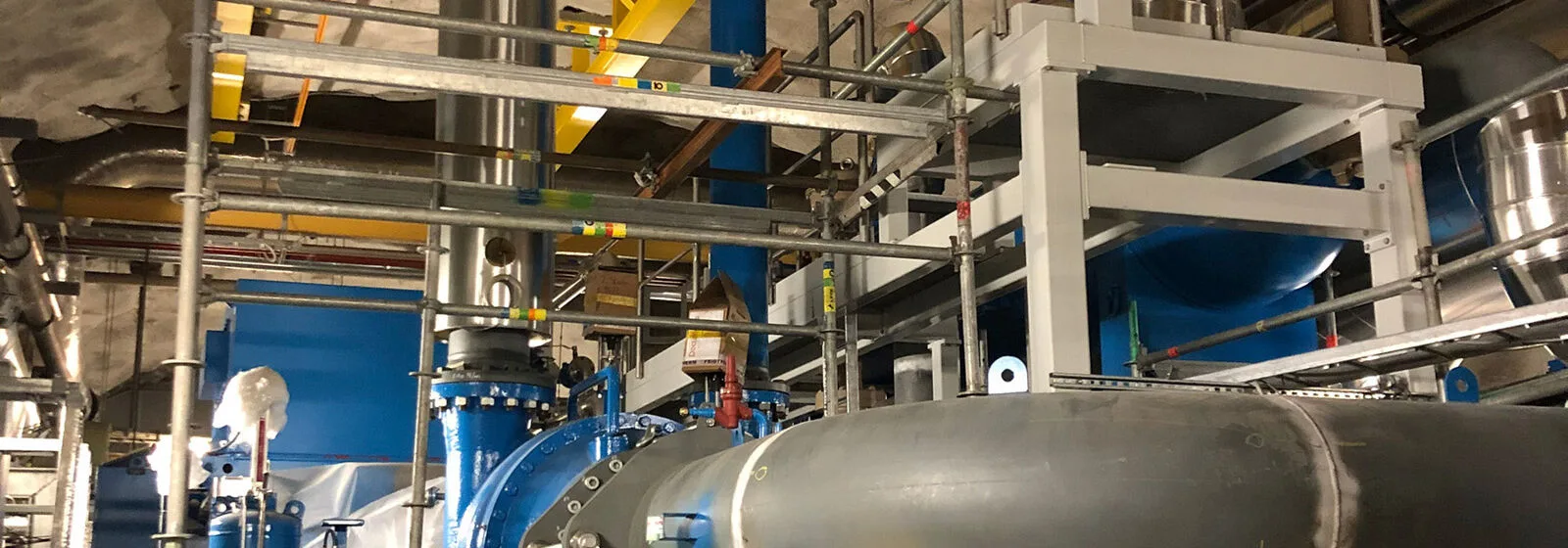 Industrial setting with large blue and metal pipes, valves, and structural supports, likely part of an energy or processing plant