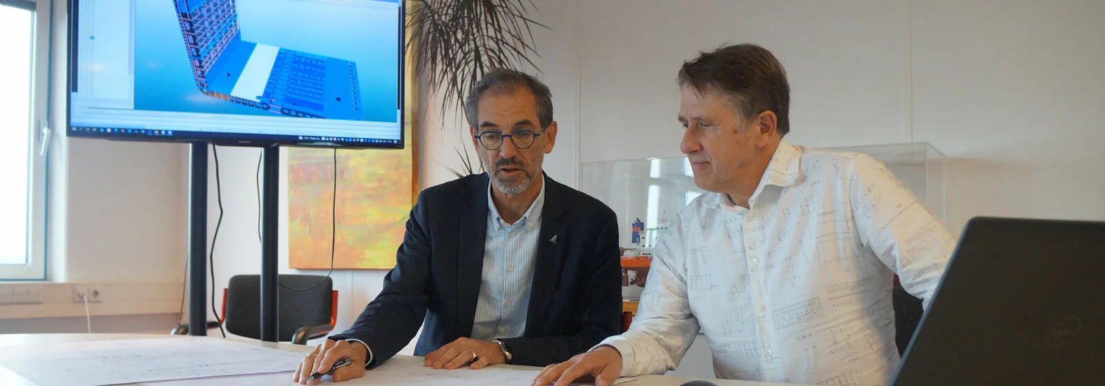 Two people reviewing documents at a table with a large screen showing a 3D ship hull model in a well-lit office setting