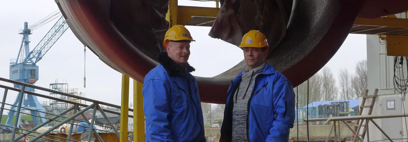 Two workers in blue jackets and yellow hard hats standing in front of industrial shipbuilding structure with scaffolding and crane