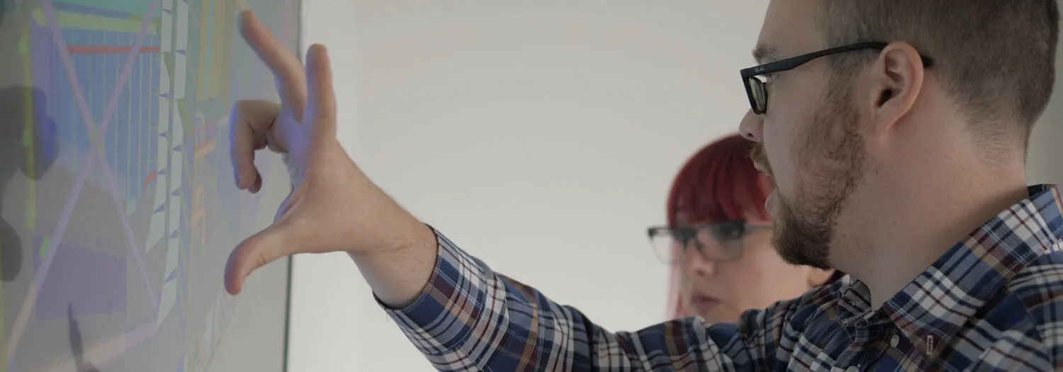 Two individuals interacting with a large touchscreen displaying a technical ship design diagram, with one person pointing at the screen.