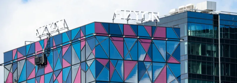 Modern building with triangular blue and pink glass panels and 'SKYON' sign on top, next to a reflective glass structure