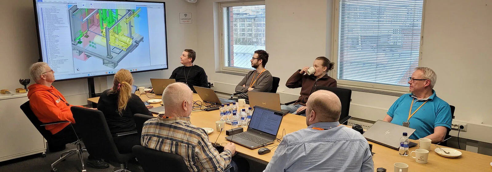 Seven people seated around a conference table viewing a 3D industrial model on a large screen, with laptops and documents in a meeting room