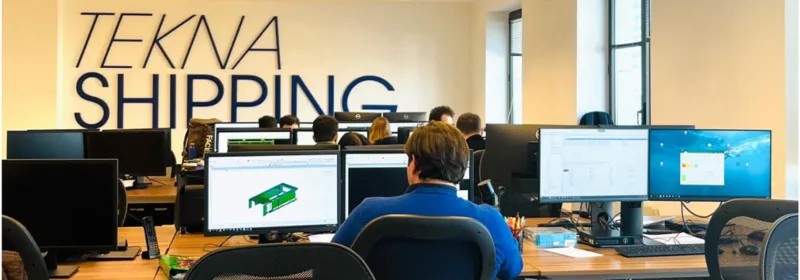 Office environment with people working at desks and computer monitors, with 'TEKNA SHIPPING' displayed on the wall in the background