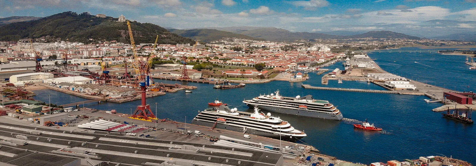 Shipyard with cranes and buildings, three expedition cruise vessels docked in the water, and a town with hills in the background
