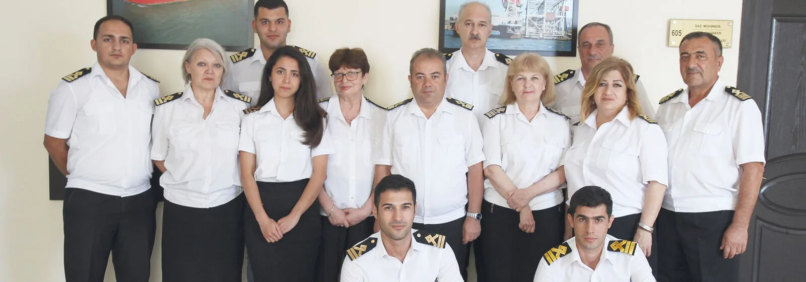 Group of maritime professionals in white uniforms with gold-striped epaulettes posing in front of framed ship photos and a labeled door