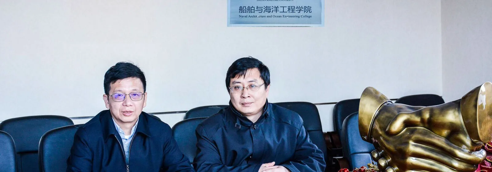 Two individuals seated at a table with a golden handshake sculpture in front, and signage for Dalian Maritime University in the background