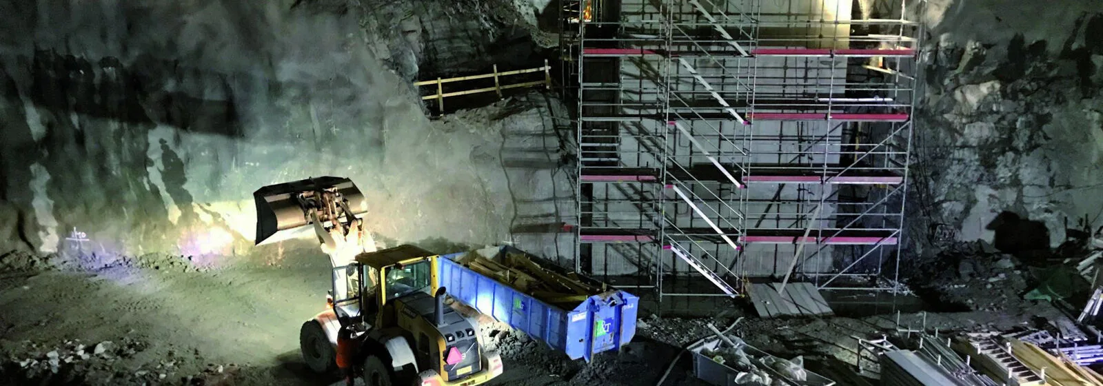 Underground construction site with excavator, blue container, scaffolding against rock wall, and scattered construction materials