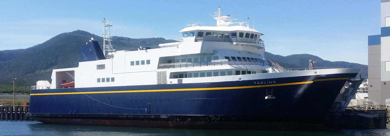 Ferry named TAZLINA with blue hull and yellow stripe docked at a port, with mountains and clear skies in the background