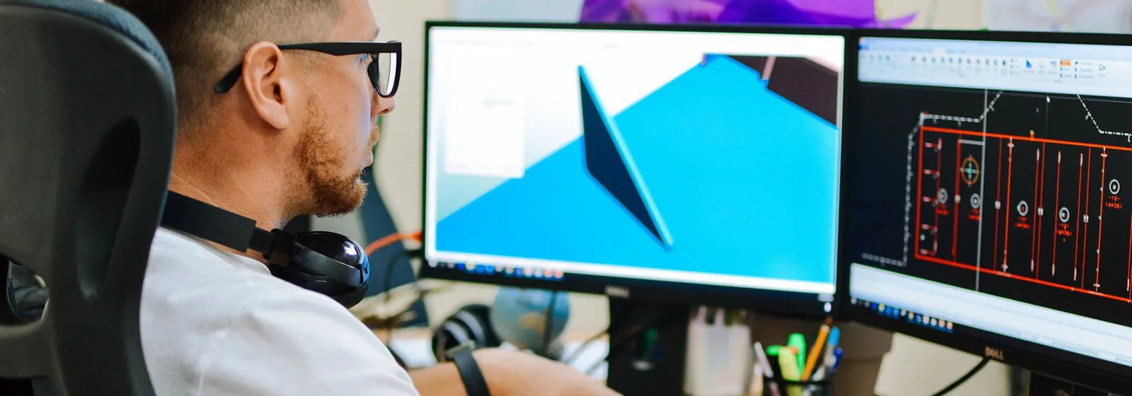 Person at desk with two monitors showing 3D ship model and technical naval architecture drawings, surrounded by office supplies