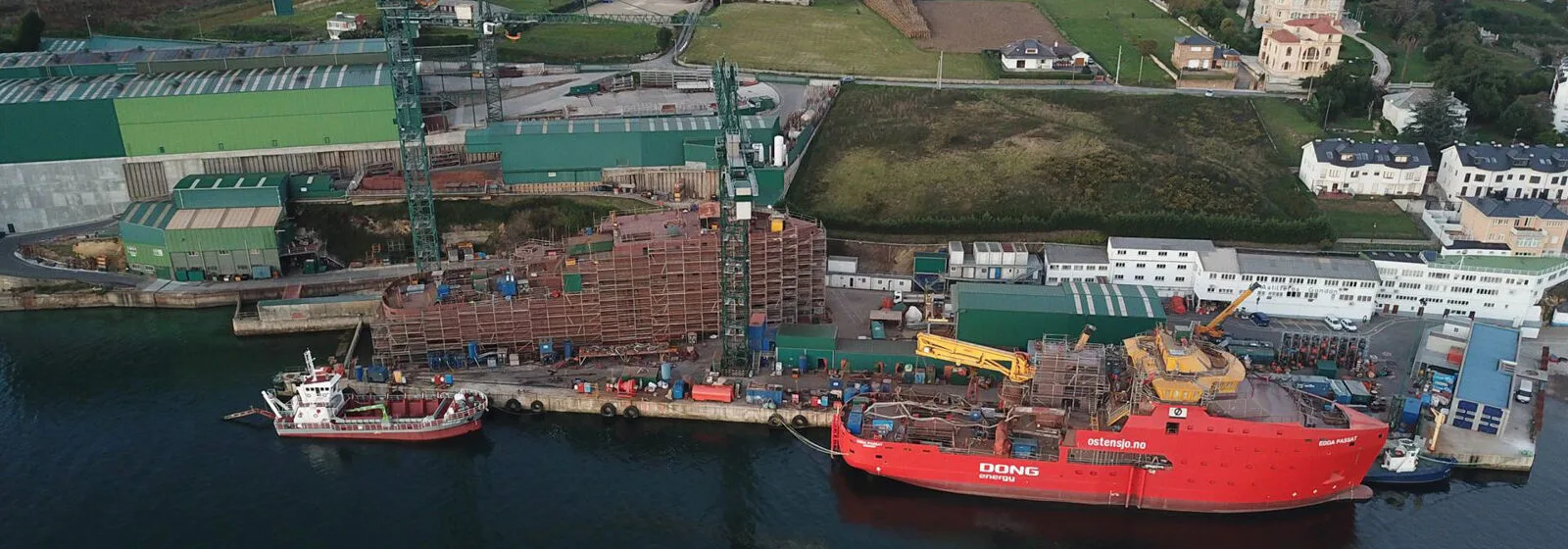 Aerial view of Gondan Shipyard with two docked ships, green buildings, cranes, and surrounding grassy and residential areas