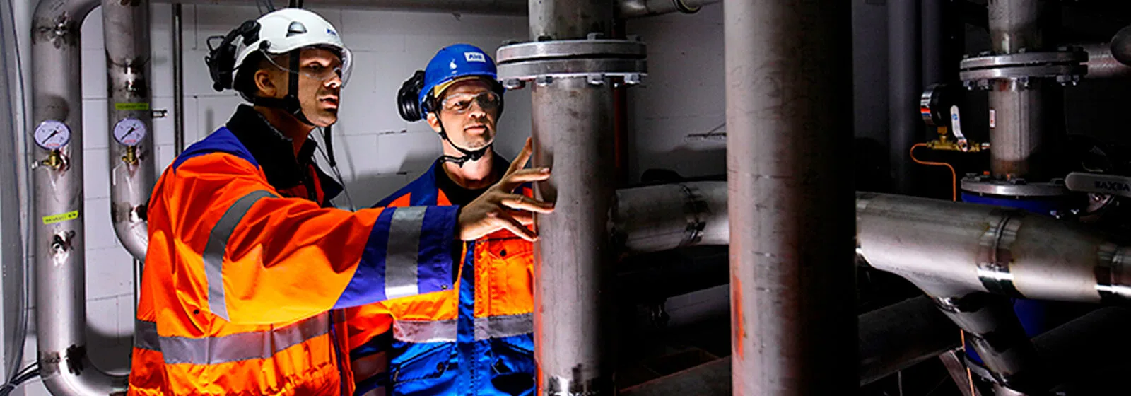 Two people in safety gear working with large metal pipes, gauges, and valves in an industrial engineering environment