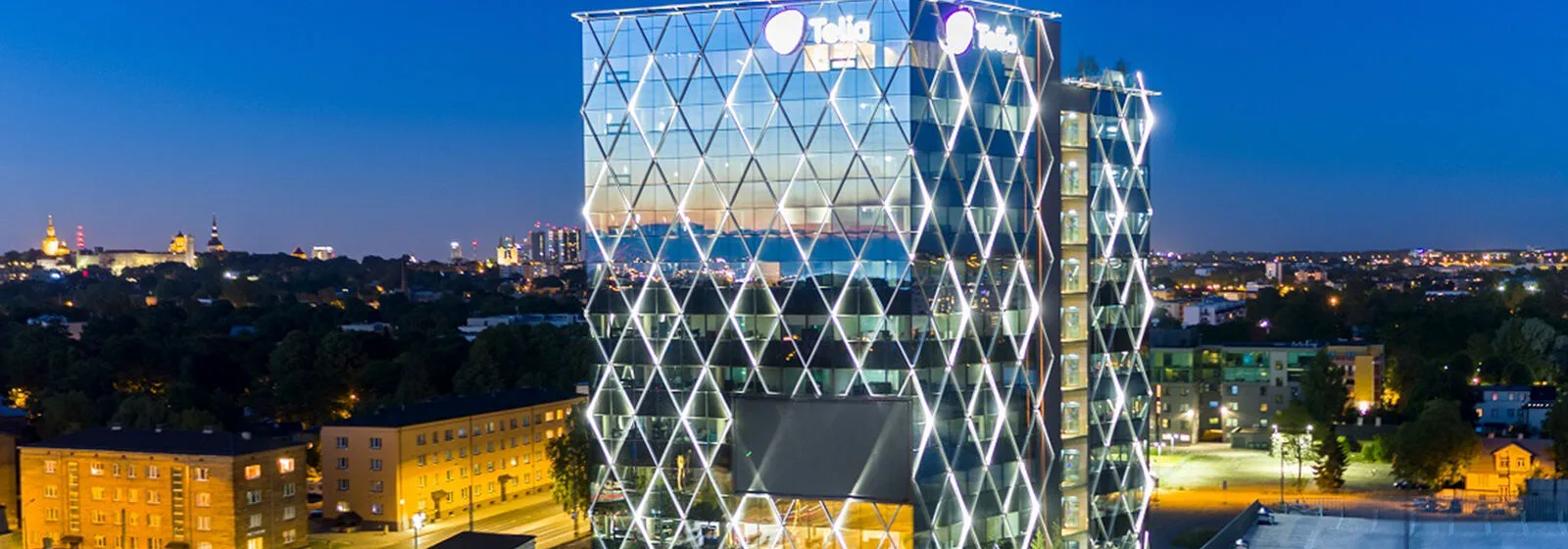 Modern office building at night with illuminated triangular facade patterns and the Telia logo at the top, surrounded by city lights