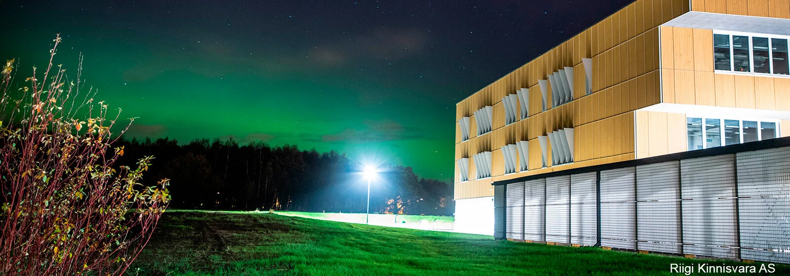 Modern wooden building with large windows near grassy field under green-lit sky, labeled Riigi Kinnisvara AS