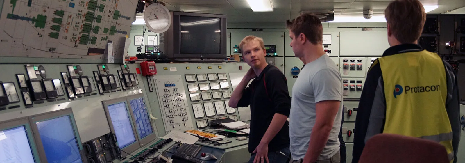 Control room with three people observing monitors and control panels, one wearing a yellow vest labeled Protacon
