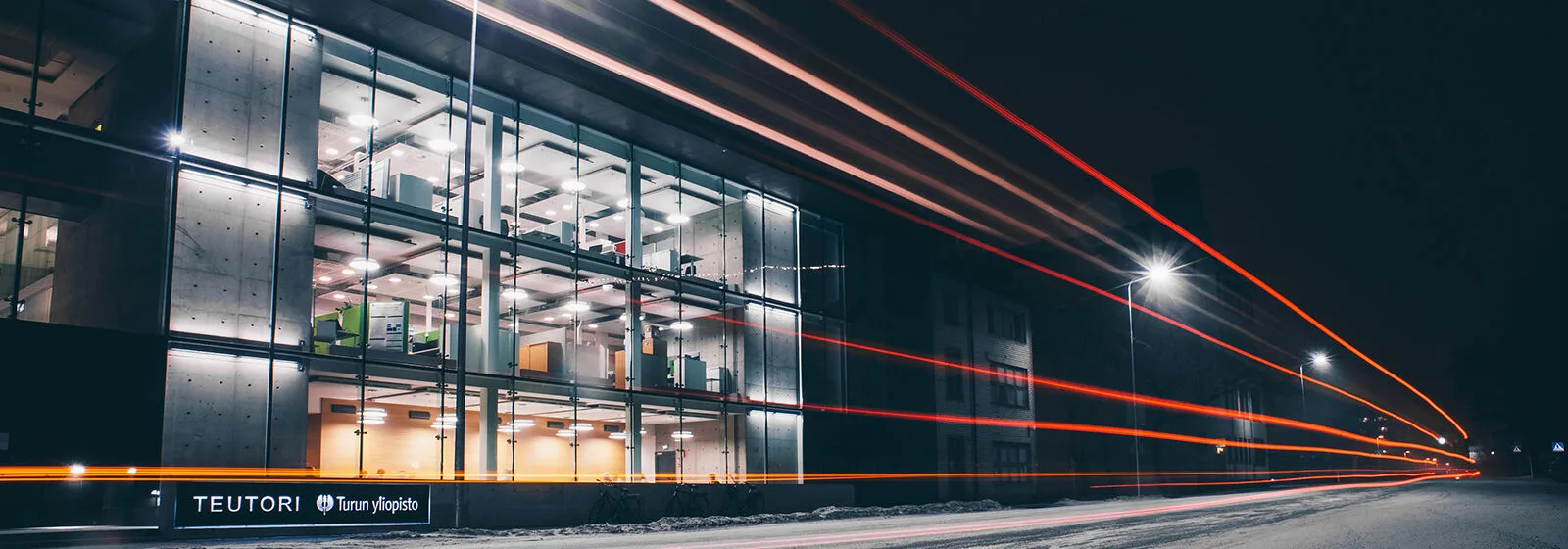 Modern glass-fronted building at night with illuminated offices, 'TEUTORI' and 'Turun yliopisto' signage, and light trails from passing vehicles