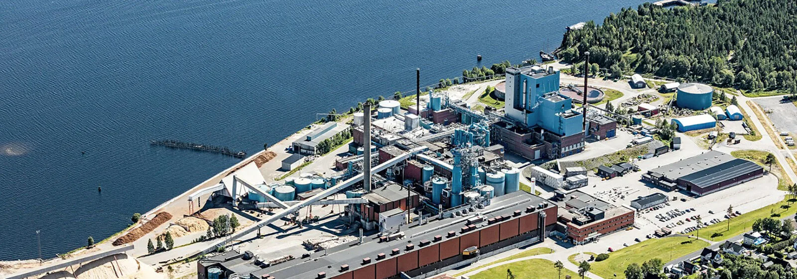 Aerial view of industrial pulp mill facility near water, with large buildings, storage tanks, chimneys, and surrounding roads