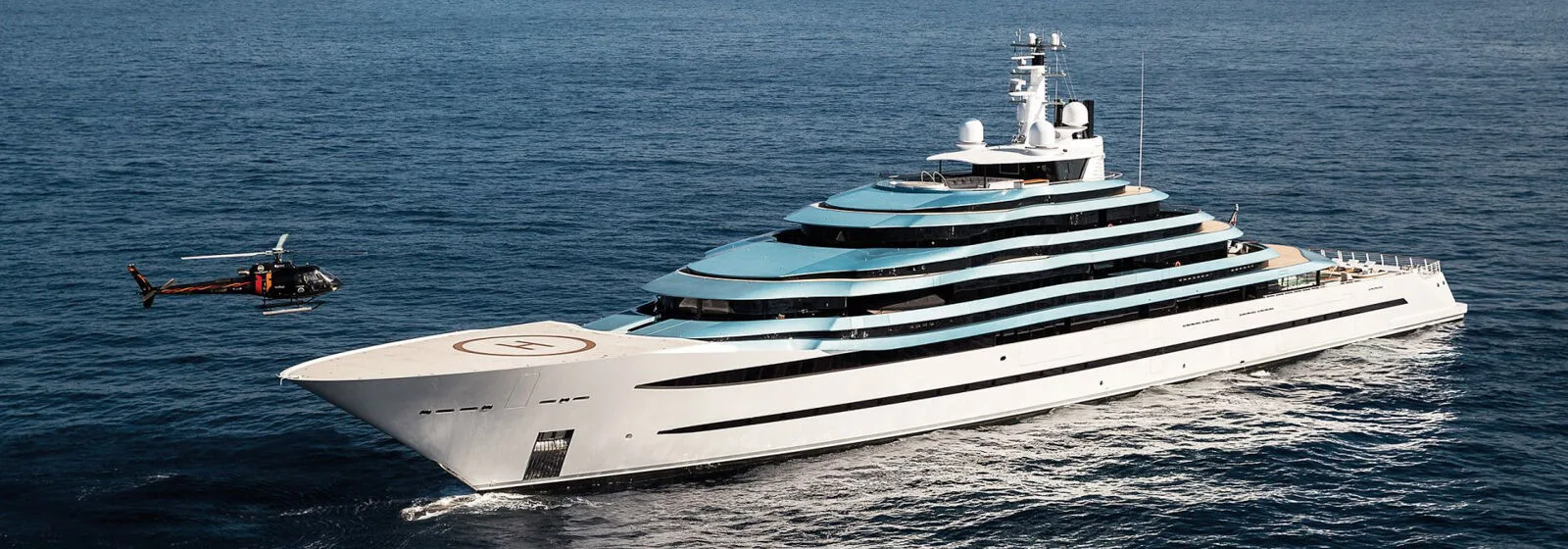 Large luxury yacht with multiple decks and helipad cruising on calm sea, with helicopter flying nearby