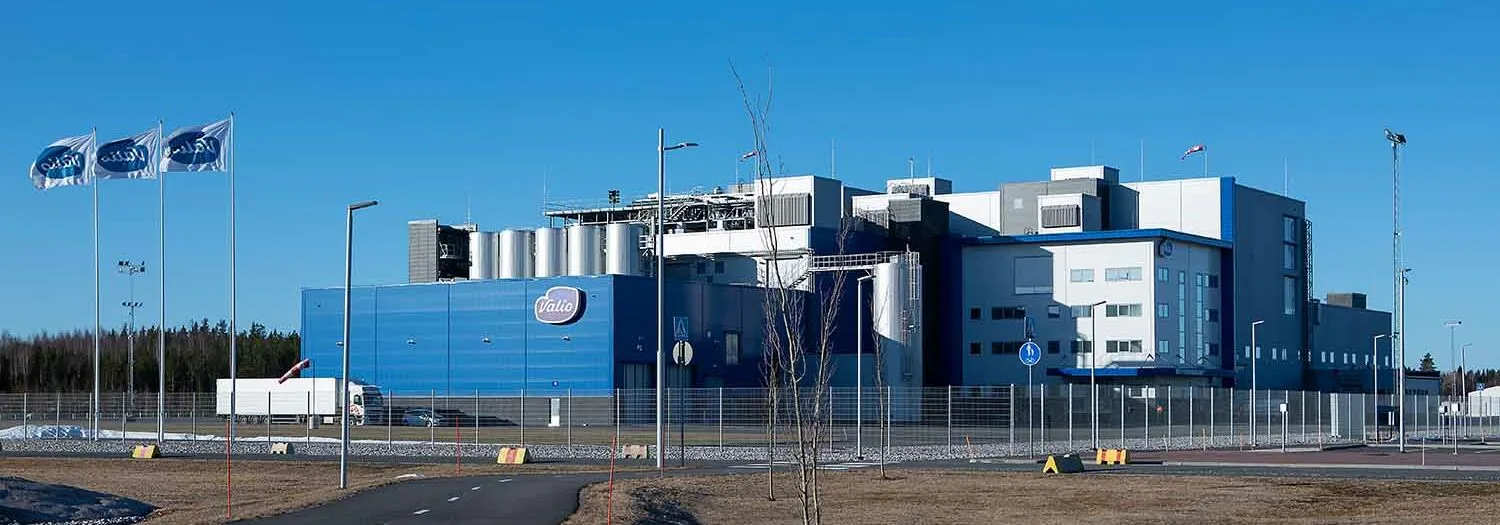Industrial facility with a blue and white building, Valio flags, cylindrical structures, and a truck parked near the fenced area