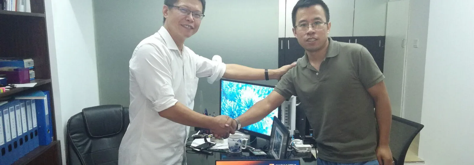 Two individuals shaking hands in an office with Cadmatic logo on a monitor, surrounded by desks, chairs, and shelves with binders