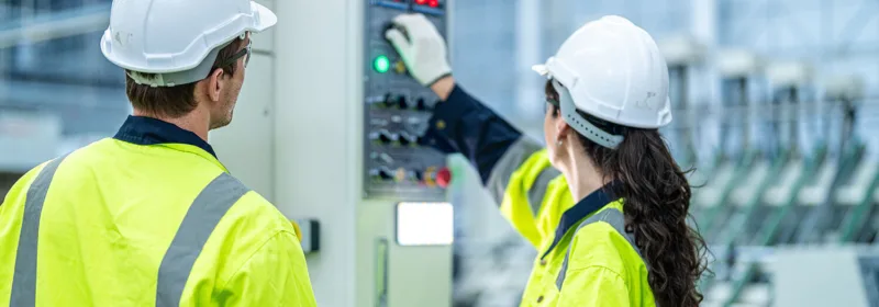 Two people in high-visibility jackets and hard hats working at an electrical control panel in an industrial environment