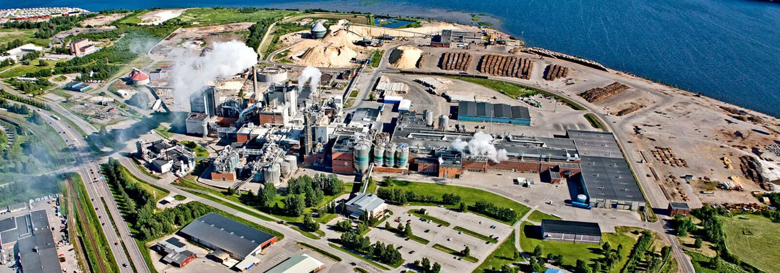 Aerial view of pulp mill facility near water with buildings, tanks, raw material piles, and steam emissions surrounded by greenery
