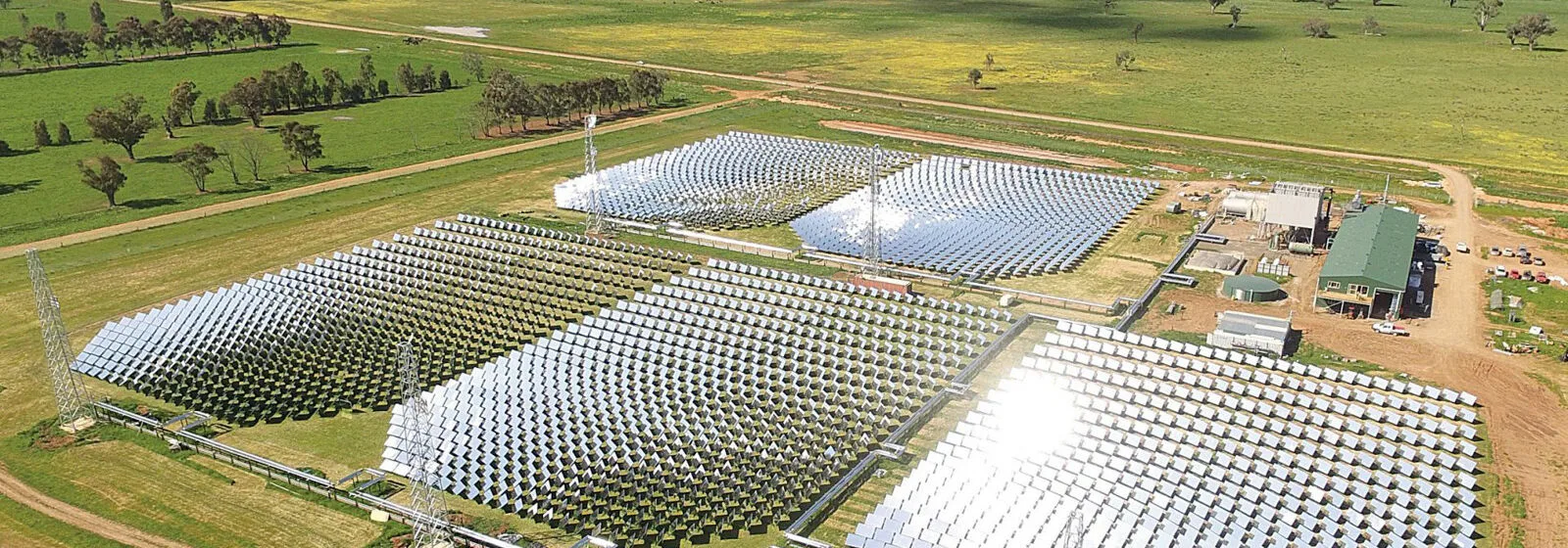 Large solar power plant with grid-arranged panels reflecting sunlight, surrounded by green fields and operational buildings
