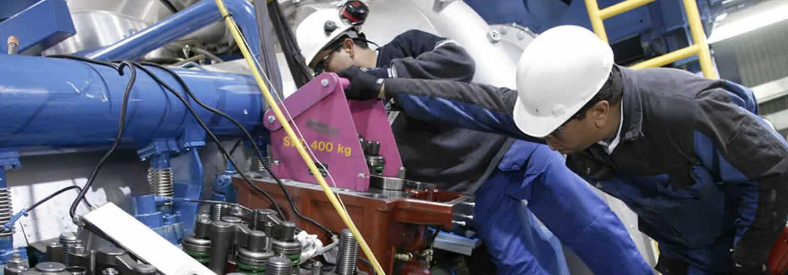 Two workers in hard hats inspecting industrial machinery with pipes, bolts, and a pink component labeled '500-400 kg
