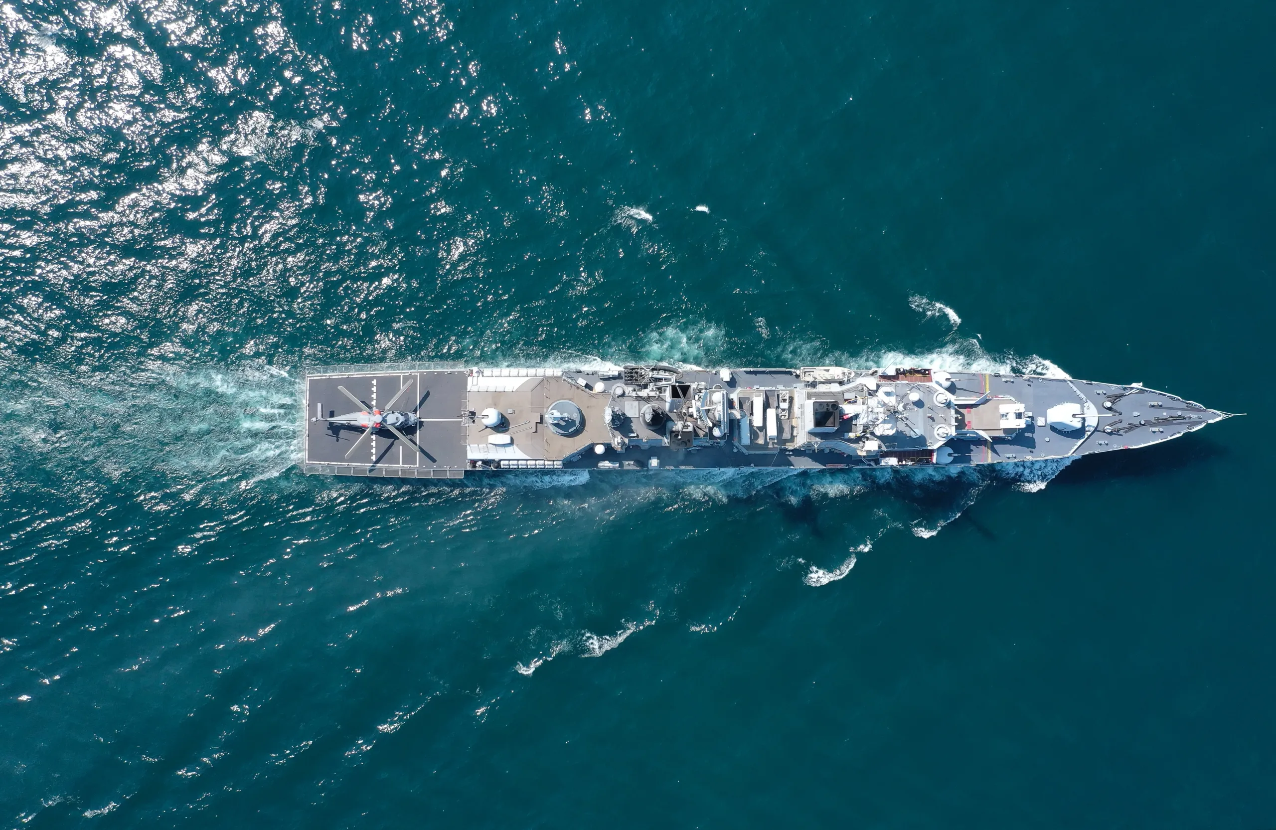 Top-down aerial view of a modern naval warship cruising through the ocean, featuring a helicopter on its rear landing pad and various radar and weapon systems on deck.